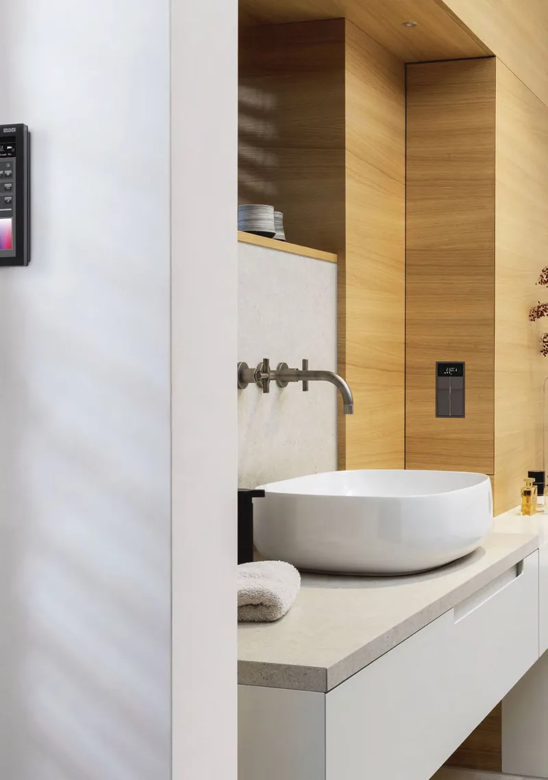Łazienka z drewnianą zabudową i umywalką nablatową, panel dotykowy Smart Control na ścianie i sterownik pomieszczenia przy lustrze — JUNG