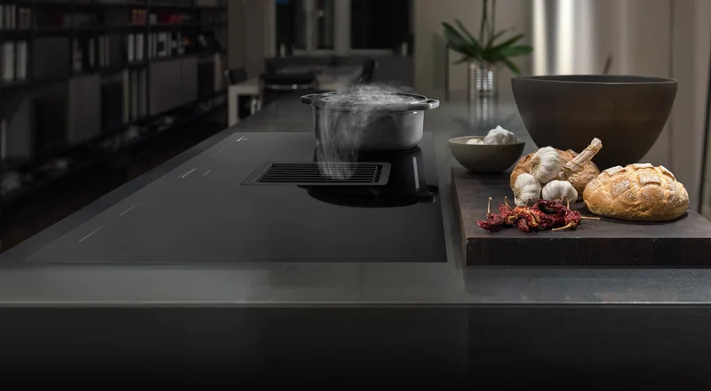 Plyta indukcyjna Smeg ze zintegrowanym okapem downdraft na ciemnej wyspie, garnek z parujaca potrawa — Smeg
